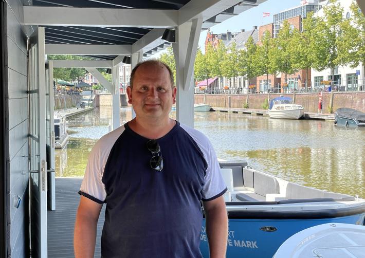 Johan poseert op de opstapboot in de haven van Breda met een blauwe sloep op de achtergrond