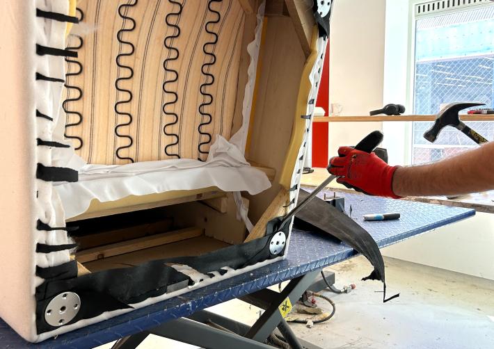 Een fauteuil ligt gekanteld op een werkbank. Iemand plaatst een schroevendraaier achter de poot en slaat er met een hamer op.